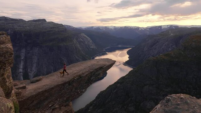 Hiker with dog stands on Trolltunga cliff in Norway at sunset, feeling awe and peace, Trolltunga