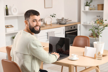 Obraz premium Young bearded man working with laptop at table in kitchen