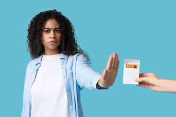 Young African-American woman rejecting pack of cigarettes on blue background. Stop smoking concept