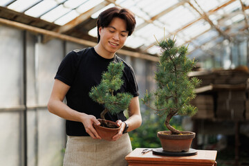 Smiling asian florist holding spruce bonsai in greenhouse