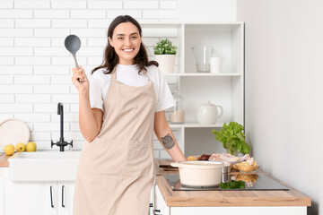 Young housewife with ladle cooking soup on stove in kitchen
