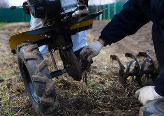 Farmer wearing gloves is diligently removing roots from the plow of a two-wheel tractor while...