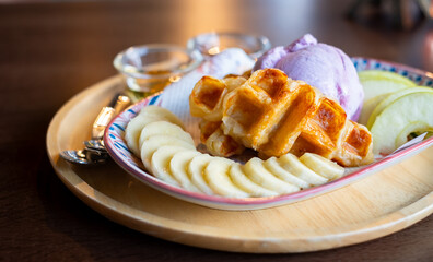 Delicious dessert plate featuring golden Belgian waffles, sliced bananas, green apples, and a scoop of purple ice cream, served with honey syrup. Perfect sweet treat presented on a wooden tray