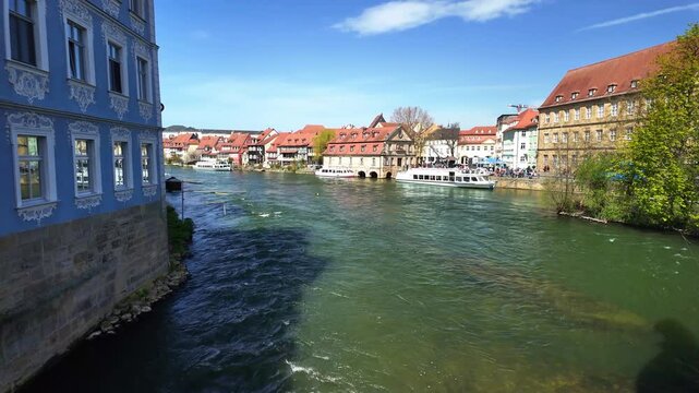 A well-lit Bright sunny day for tourism with blue sky and flowing water in the foreground and beautiful architecture and tourists on boats in the background in Bamberg Bayern Bavaria Germany Europe