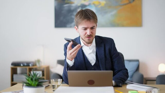 A businessman is using the voice command feature on his phone while working on his laptop. He's likely multitasking to stay productive.