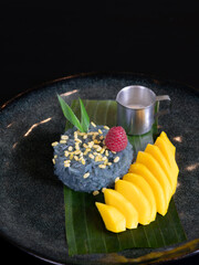 A plate of ripe mango with sticky rice and coconut milk sauce.  Various sweet Thai desserts in background with Thai style plate decorations such as green banana leaves. 