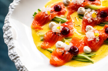 A gourmet dish featuring thin slices of salmon arranged with cucumber, garnished with berries and dollops of cream, all presented on a vibrant sauce.