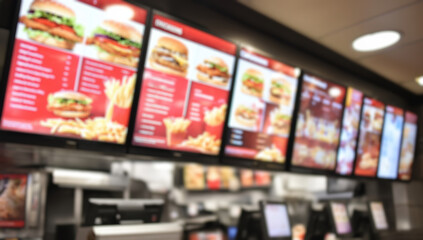 A blurred background of a fast food restaurant interior, with digital menu boards displaying meal options and a customer sitting at a table, using a mobile phone to order online.