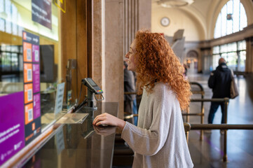 Young woman buying train ticket at ticket office window in train station