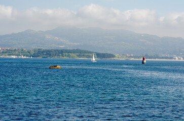 Obraz premium Peaceful view of a single sailboat gliding on calm blue water with a distant coastline and relaxing mountains under a cloudy sky. Tranquil maritime travel scenery.