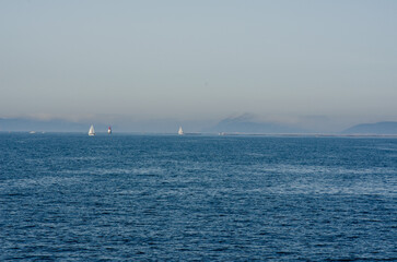 Peaceful seascape featuring sailing boats gliding across calm blue water, with a lighthouse and hazy mountain range on the horizon. Ideal for themes of serenity and travel.