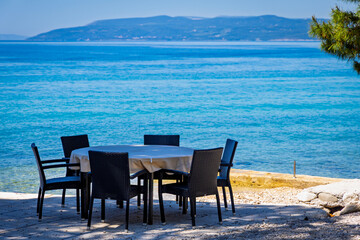 Empty seaside restaurant table with six chairs by the turquoise Adriatic Sea. Idyllic Mediterranean setting for outdoor dining with scenic coastal views.