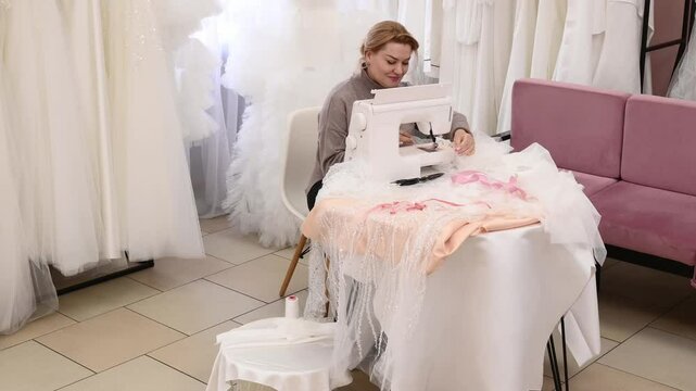A woman seamstress in a wedding salon sews a bride's dress and veil at a table and with an overlock sewing machine. Handmade, custom tailoring