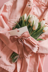 White tulips wrapped in a soft pink denim jacket with a satin pink hair bow on a card, styled on a pastel pink background — a charming and delicate flatlay for spring or celebration