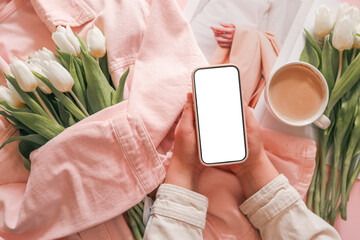 Woman holding smartphone with blank screen, white tulips in pink denim jacket, coffee and fashion...