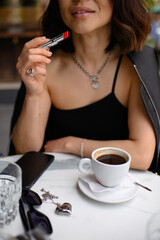 Asian woman sitting at an outdoor café table, applying red lipstick. A cup of coffee in front of her. Stylish, candid moment of beauty and daily routine in urban setting.