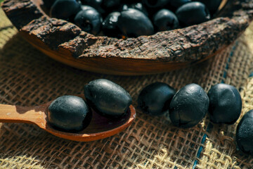 Rustic wooden bowl filled with black olives and a wooden spoon on burlap fabric, captured in warm natural light for a Mediterranean kitchen or food concept