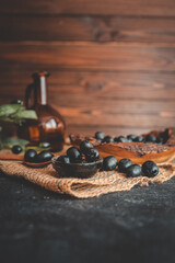 Rustic still life composition with black olives in a wooden bowl, a wooden spoon, olive branch, and glass bottle on burlap and dark table. Warm vintage food photography style