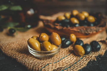 Rustic composition of green and black olives in a glass bowl with olive branches, burlap cloth, and a wooden bowl in warm natural light on a dark textured background