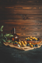 Rustic composition of green and black olives in a glass bowl with olive branches, burlap cloth, and a wooden bowl in warm natural light on a dark textured background