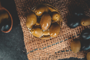 Top view of green olives in a glass bowl on rustic burlap with scattered black olives and wooden...