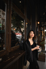 Asian woman standing alone by a café bar counter on a city street, enjoying her morning coffee and quietly observing the urban scenery before starting her workday.
