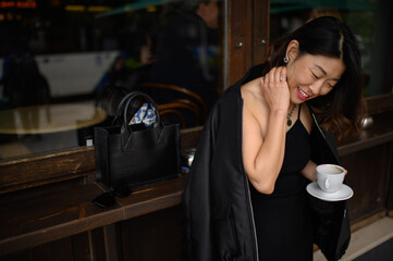 Asian woman standing alone by a café bar counter on a city street, enjoying her morning coffee and quietly observing the urban scenery before starting her workday.
