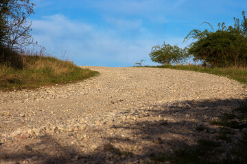 An empty gravel road winds uphill through a rural landscape. The path leads toward the horizon under a blue sky with soft clouds