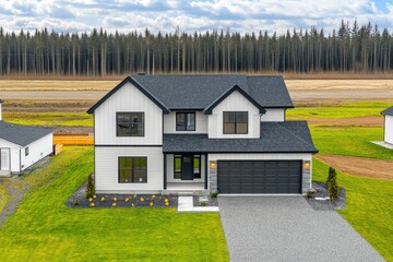 Obraz premium Modern farmhouse style home with white siding, dark gray roof, and black garage door. Situated on a grassy lot, amidst a forest backdrop