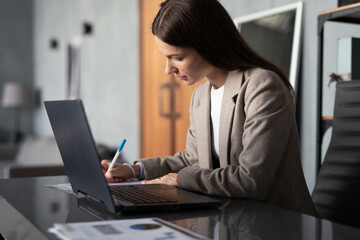 Young woman sitting at desk in office, working on her laptop and writing notes documents home office