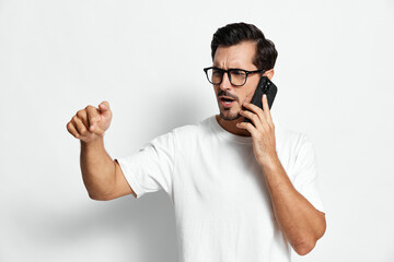 Serious young man talking on mobile phone, wearing white t-shirt and glasses, expressing concern, gesturing while interacting isolated on plain background. Communication concept.