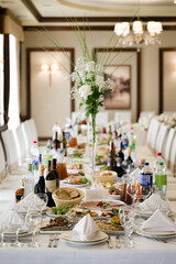 A long table is elegantly set for a banquet. White flowers and glassware adorn the table, alongside an array of food platters.