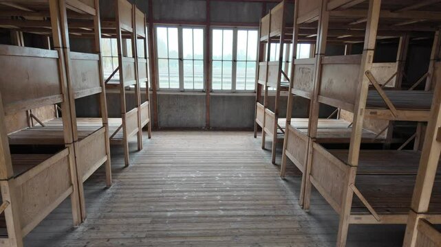 An empty wooden dormitory room with multiple rows of stacked bunk beds and a wide central aisle, illuminated by natural light from large windows in a preserved historic structure.