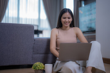 Fototapeta premium Happy freelancer working from home, sitting on the floor with legs crossed, using laptop and smiling, with a cup of coffee and a plant next to her, sofa and tv in the background