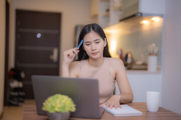 Concentrating on her work, a young Asian woman sits at a wooden table in a cozy kitchen, using her...