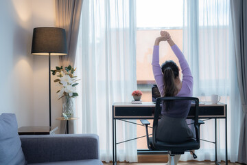 Young Asian businesswoman sitting on an ergonomic chair is stretching her arms after using a laptop computer for a long time in her modern home office