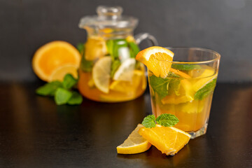 Orange and mint lemonade in a clear glass with orange slices in front on a blurred dark background with a teapot of orange drink behind. Natural refreshment, diet drinks, healthy eating.