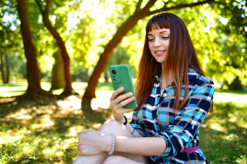 A young woman in dress with long hair sitting on the grass in the nature looking at her phone and smiling.