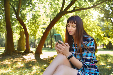 Charming lady in a dress, sitting on the green grass in nature, against the backdrop of a summer park, chatting at her phone.