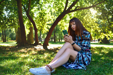 A young lady in a dress, sitting outdoors on the green grass, against the backdrop of a summer park, looking at her phone.