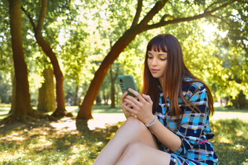 Charming lady in a dress, sitting on the green grass in nature, against the backdrop of a summer park, chatting at her phone.