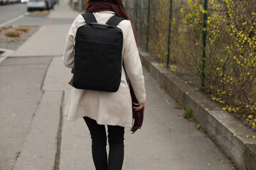 A woman carrying a black backpack is walking along a sidewalk, moving purposefully as she enjoys...