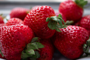 Fresh strawberries, rich red, fill a white plate, highlighted by natural light, green leafy tops contrast with the vibrant fruit. Known for high vitamin C content