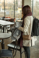 A woman is standing inside a restaurant while holding a backpack tightly in her hands, appearing deep in thought and contemplation