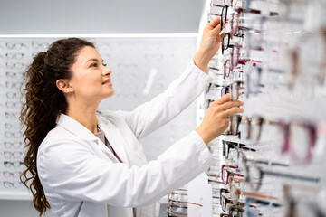 Beautiful female ophthalmologist arranging eyeglasses on the shelf in optician store.