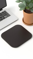 A black square mousepad sits on a white desk with a laptop and a potted plant