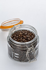 Glass jar filled with roasted coffee beans sits open on white surface, showcasing rich, dark texture of beans. jar lid is orange and slightly ajar, adding pop of color