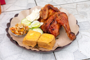 A quarter portion of crispy, golden-brown Ayam Bakar (grilled chicken) served with fried tofu, cucumber slices, sambal, and cabbage on a brown plate. Savory Indonesian meal.