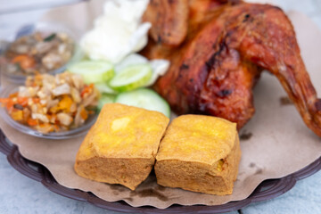 Close-up of two golden-brown Tahu Goreng (fried tofu), part of a dish with fried chicken, cucumber, sambal, and cabbage. Savory Indonesian meal.