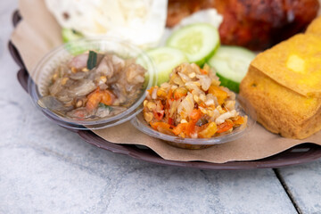 Close-up of two small bowls of vibrant sambal (chili sauce), one red and chunky, the other translucent with shallots, alongside cucumber and tofu. Spicy Indonesian condiments.